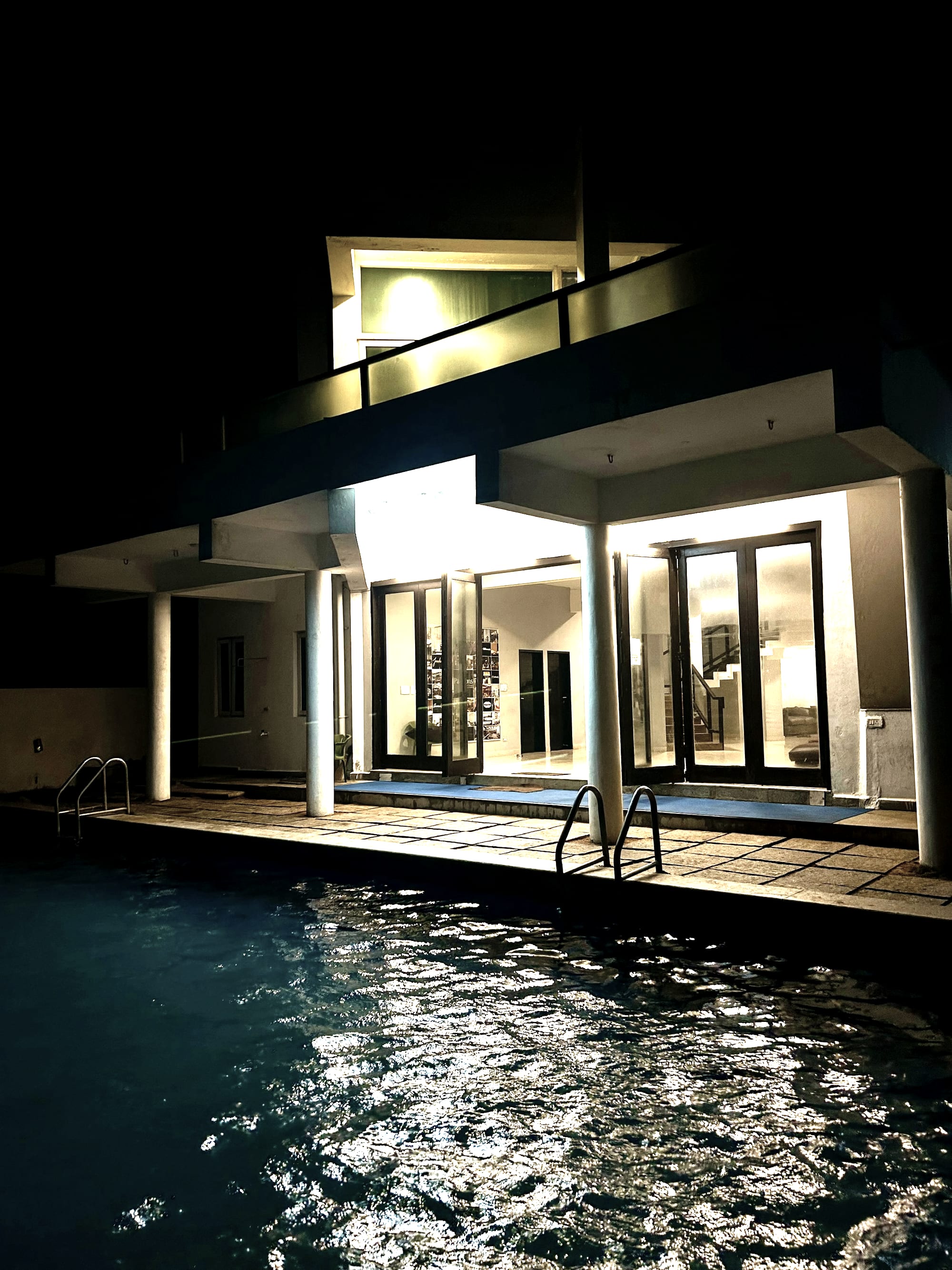 A modern house at night, the ground floor lit behind glazing, pool in front.